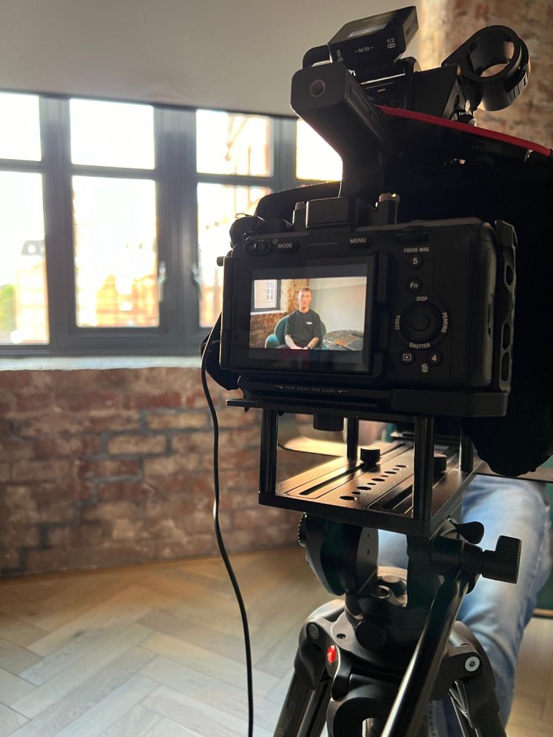 Sony camera on a tripod framing Jonny mid-workshop in the 8i Studio film room — brick wall, herringbone floor, natural light.